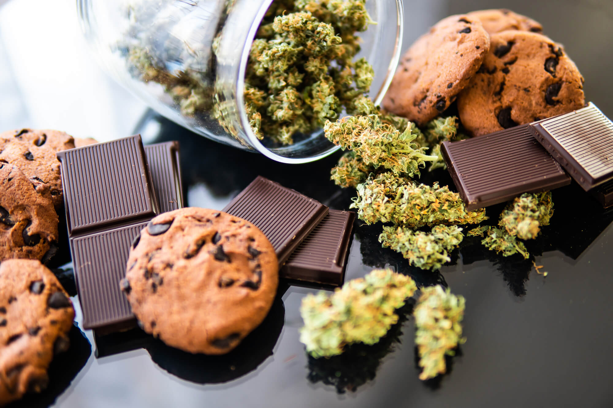 Chocolate bars, chocolate chip cookies, and cannabis buds are displayed on a black surface, with cannabis spilling out of a glass jar.